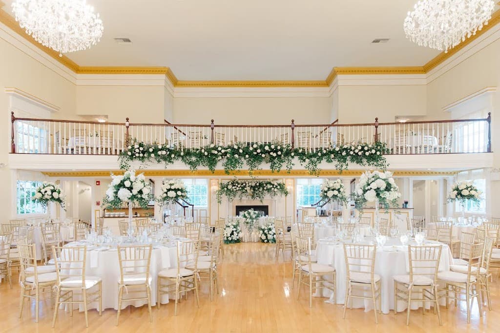 Grand ballroom wide view — both crystal chandeliers, balcony with garland, gold Chiavari chairs, tall centerpieces at The Commons 1854